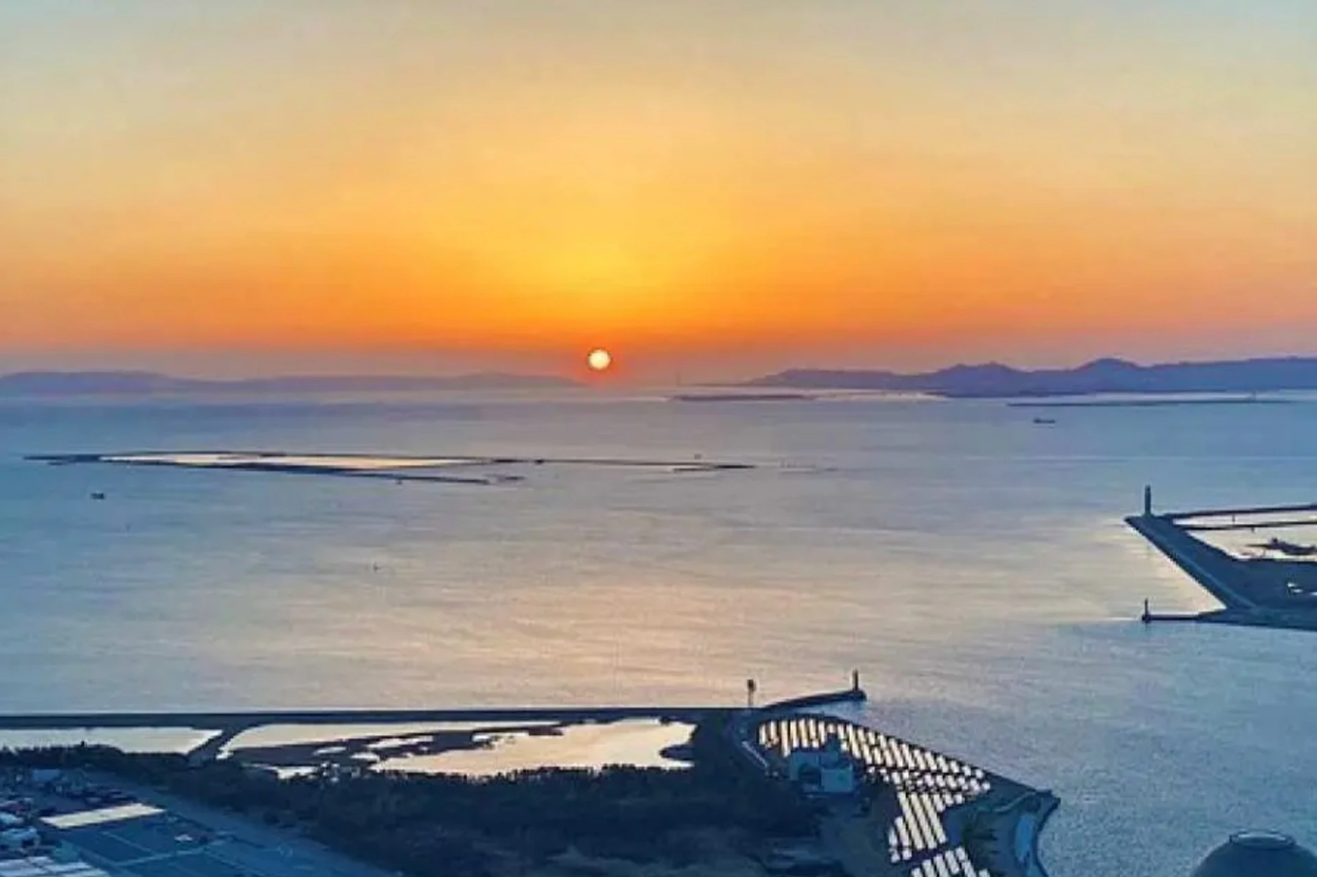 A serene coastal scene at sunset with the sun near the horizon, calm waters, and a shoreline with buildings and structures.
