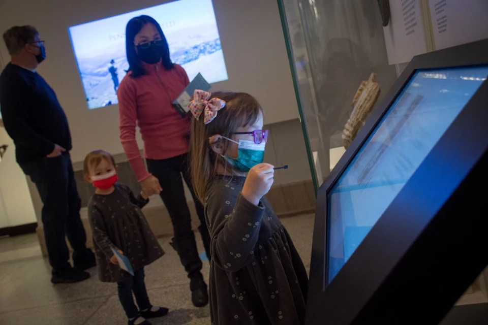 A young girl interacts with a touchscreen display in a museum while two other people, all wearing masks, observe in the background.