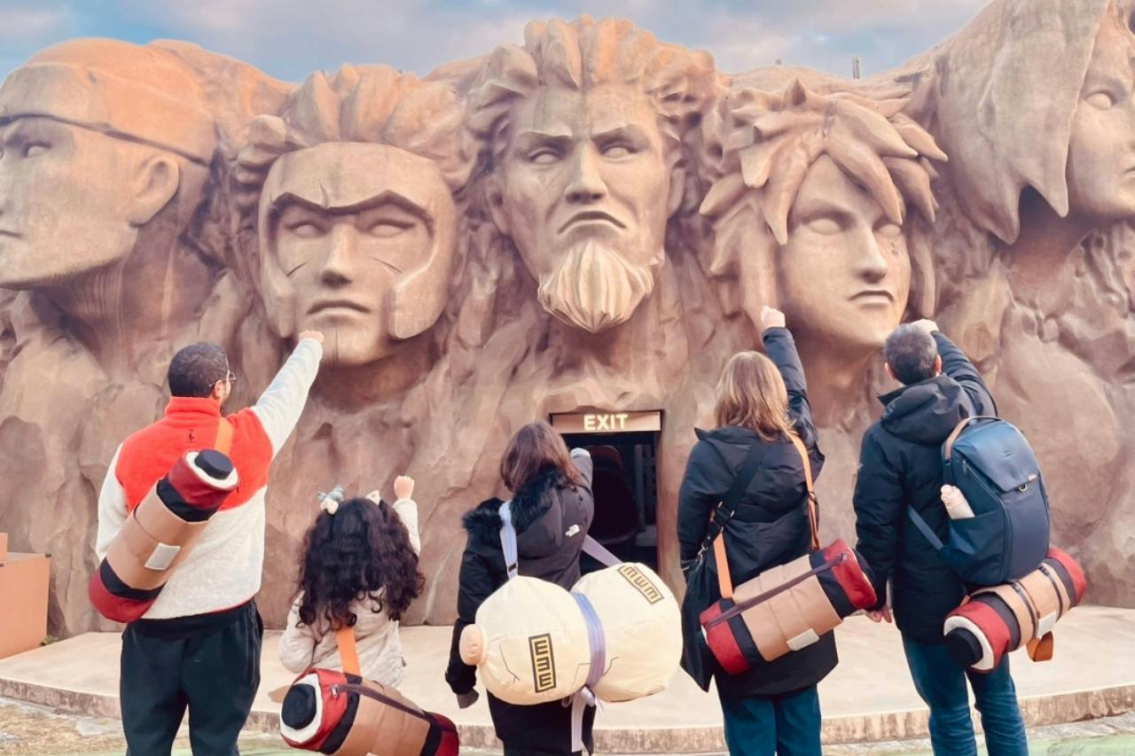 A group of people facing a large stone relief featuring four stylized faces, with an exit sign visible in the background.