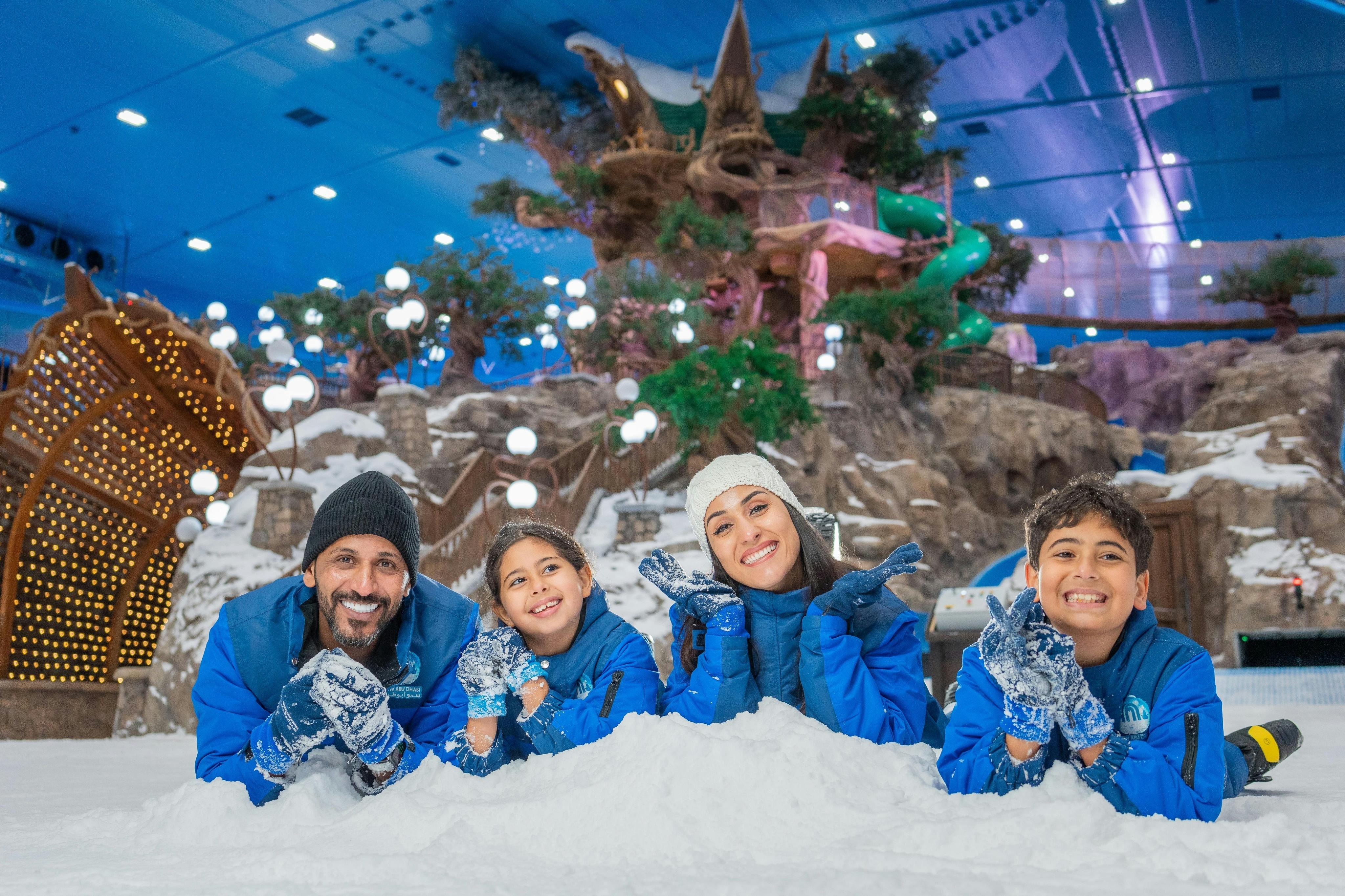 Una famiglia di quattro persone in tenuta invernale si sdraia sulla neve in un parco interno decorato, sorridendo e posando per la macchina fotografica.