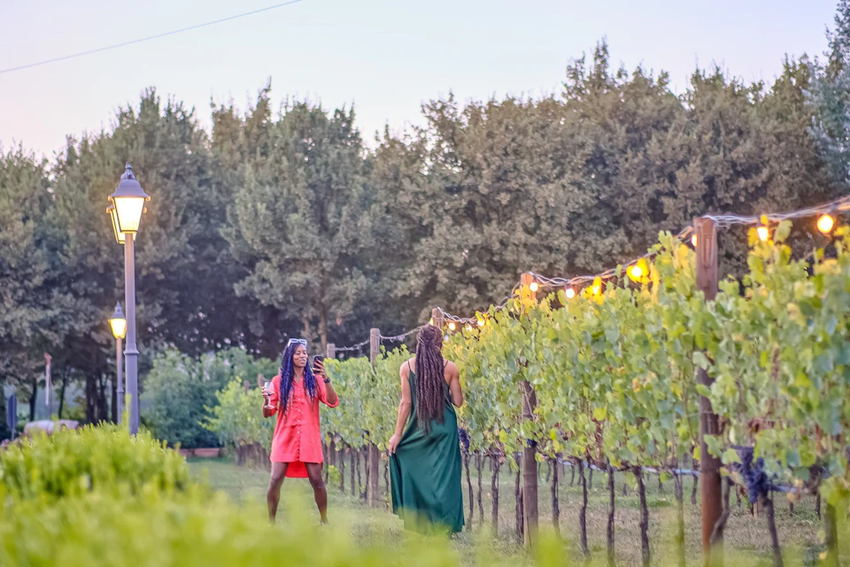 Two women walking through a vineyard, one in a red dress holding a drink, the other in a green dress walking ahead.