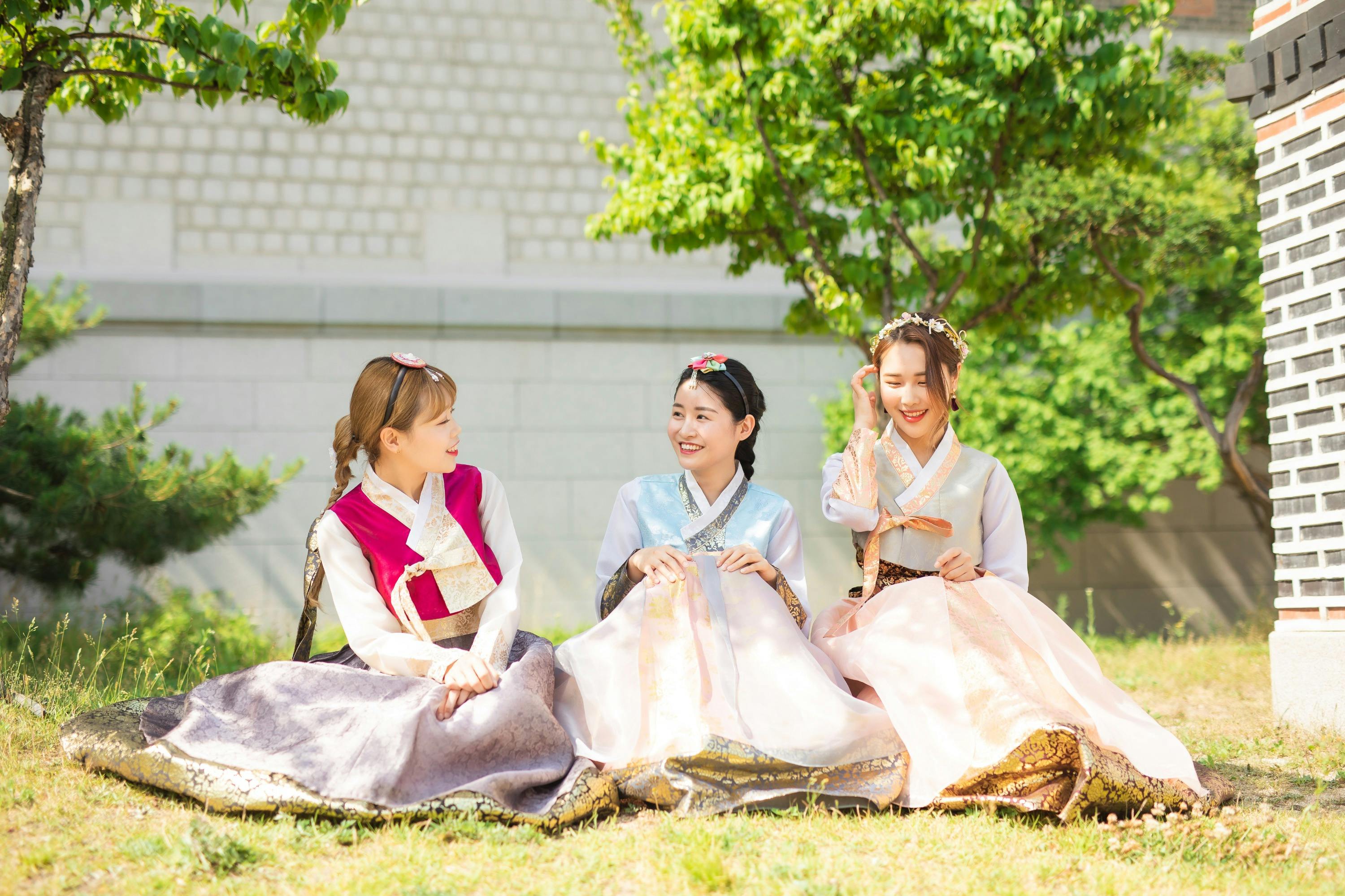 Three women in traditional Korean hanbok seated outdoors on grass, smiling, with trees and a wall in the background.