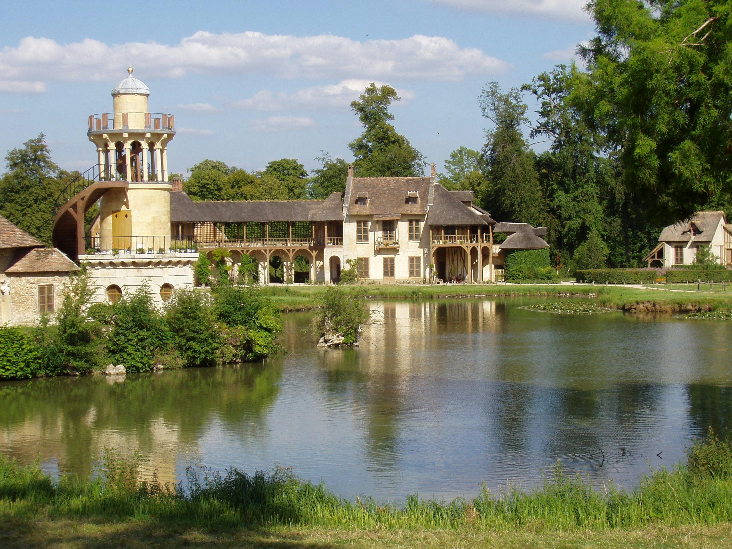 A lakeside chateau with a tower, wooden arches, lush greenery, and a reflecting water surface under a partly cloudy sky.