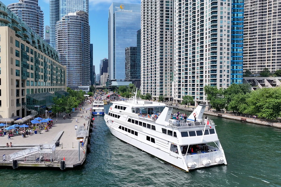 A white tour boat travels along a waterway surrounded by tall buildings and a dock with people along the side.