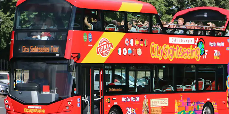 Bus touristique rouge à impériale portant la marque "CitySightseeing" et touristes sur le pont supérieur ouvert, avec des arbres en arrière-plan.