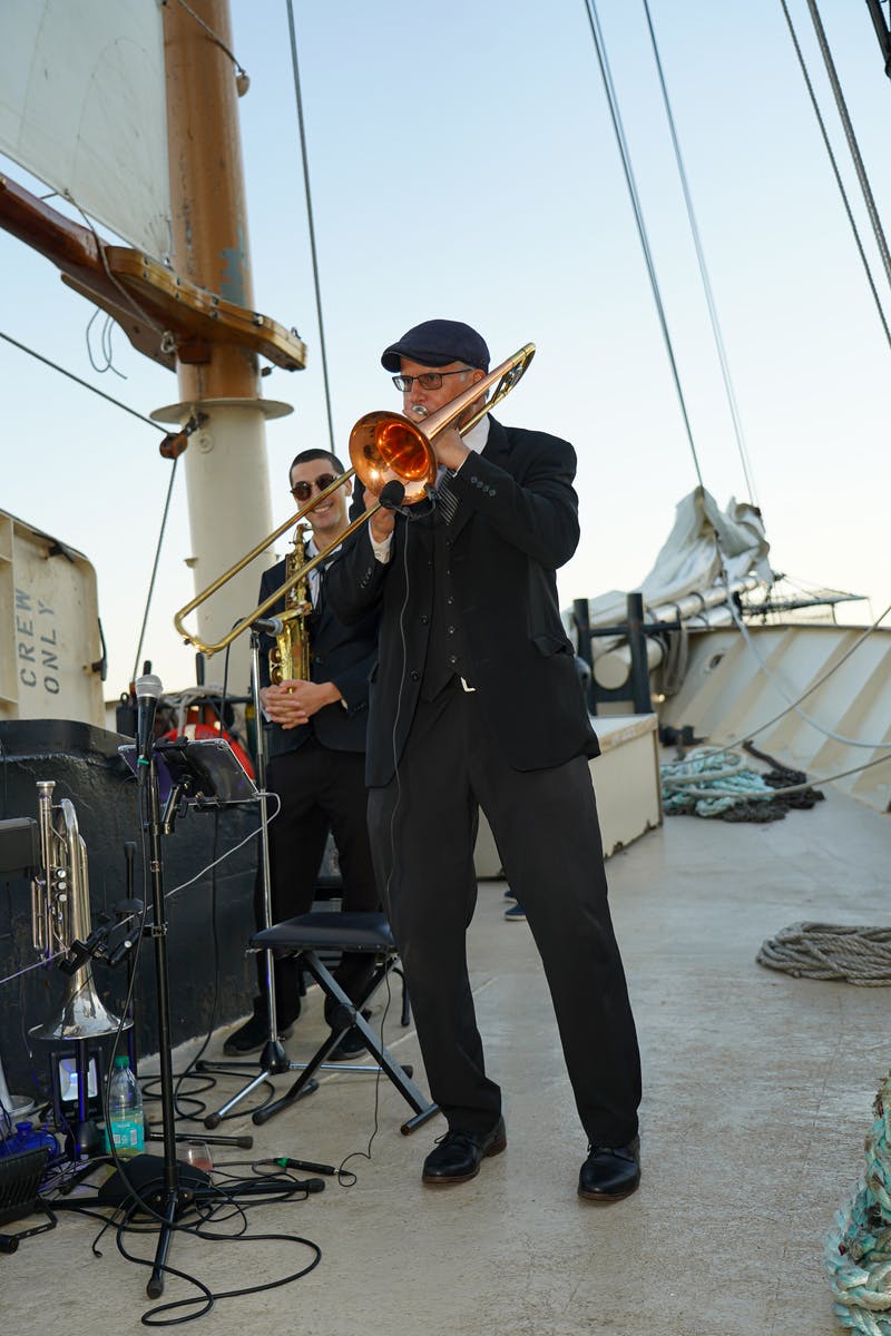 Δύο μουσικοί παίζουν χάλκινα όργανα στο κατάστρωμα ενός ιστιοφόρου πλοίου, με πανιά και σχοινιά ορατά στο παρασκήνιο.