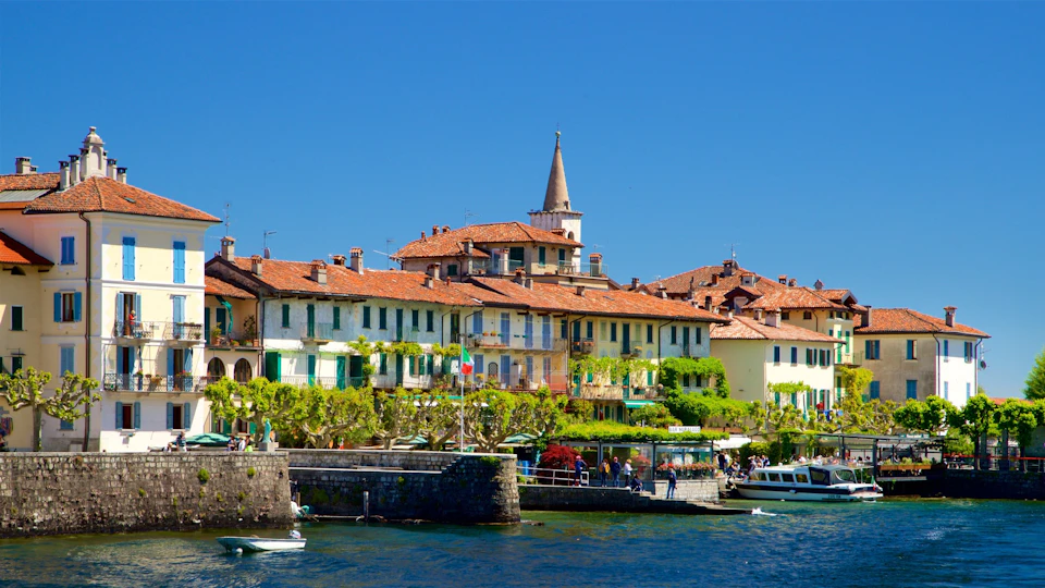 Colorful buildings with red-tiled roofs and a spire by a waterfront. People and boats are visible near the shore under a clear blue sky.