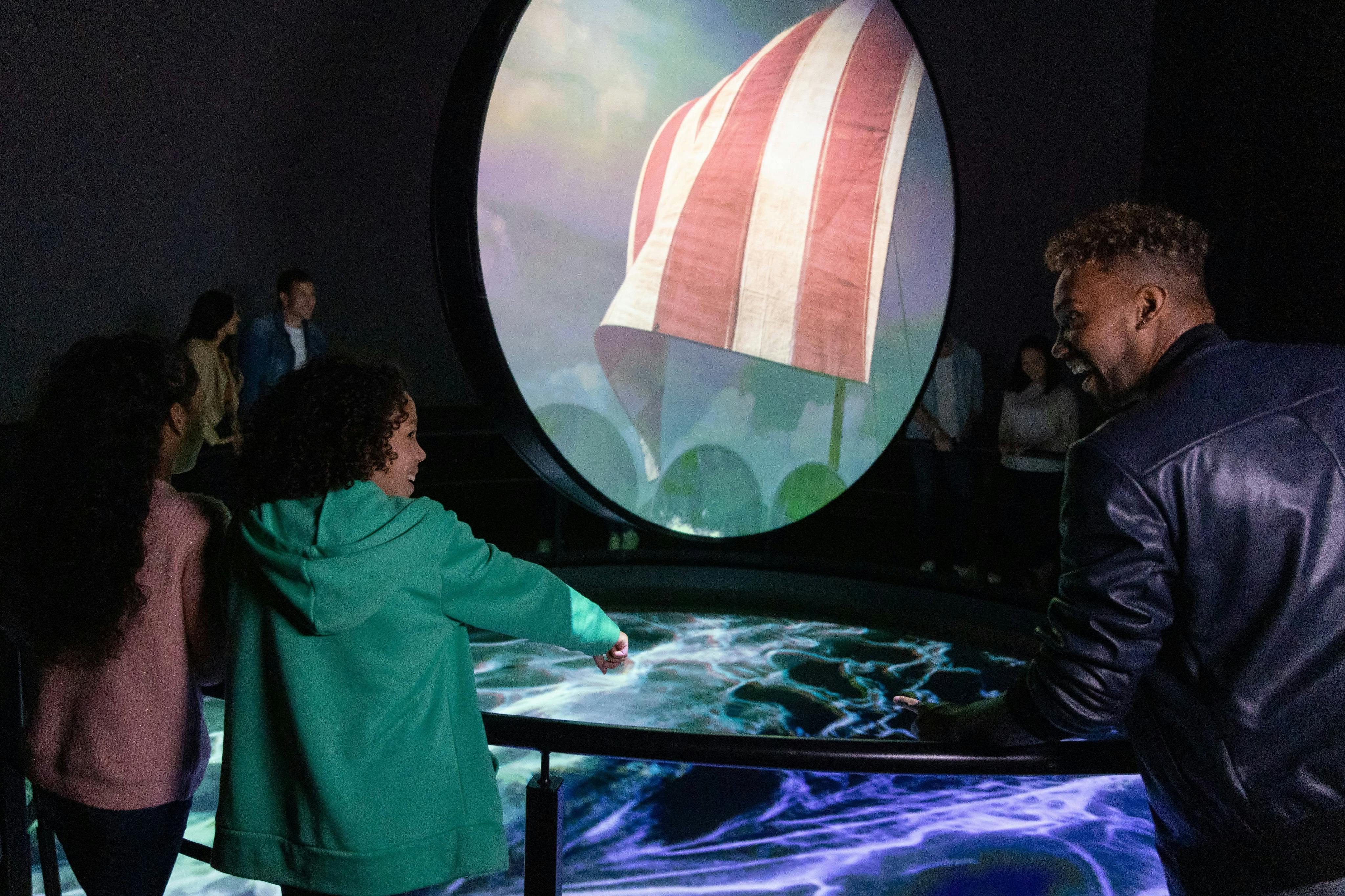 Eine Gruppe von Menschen beobachtet ein kreisförmiges interaktives Display mit wirbelnden Lichtmustern und einem großen Bildschirm, der eine gestreifte Flagge zeigt.