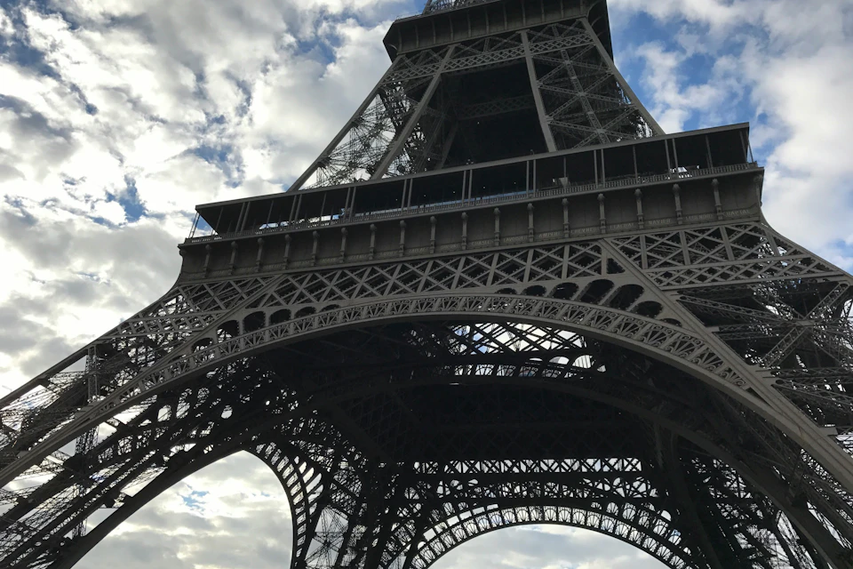 Close-up view of the Eiffel Tower from below, showing its intricate iron lattice structure against a partly cloudy sky.