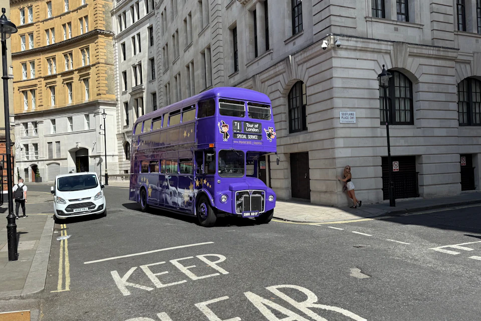 A purple double-decker bus on a city street near tall buildings. A person is standing by a wall on the sidewalk.