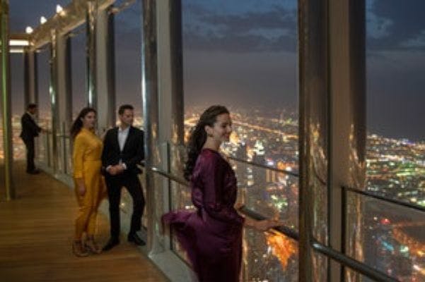 Drie mensen in elegante kleding staan &#39;s nachts op een hoog uitkijkplatform en kijken uit over een helder verlicht stadslandschap.