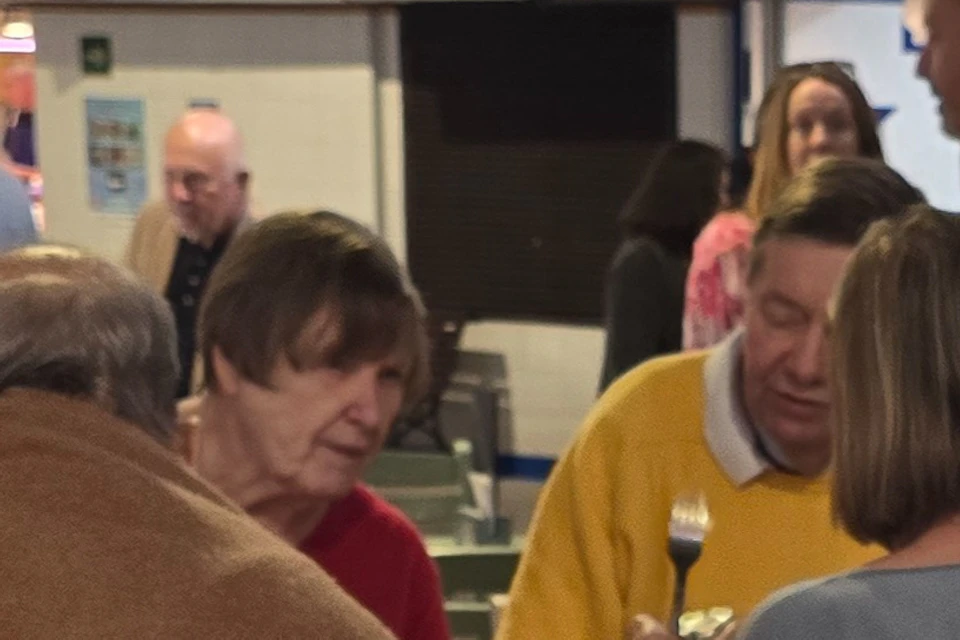 Woman smiling at a table with small dishes of food, a drink, and people conversing in the background.