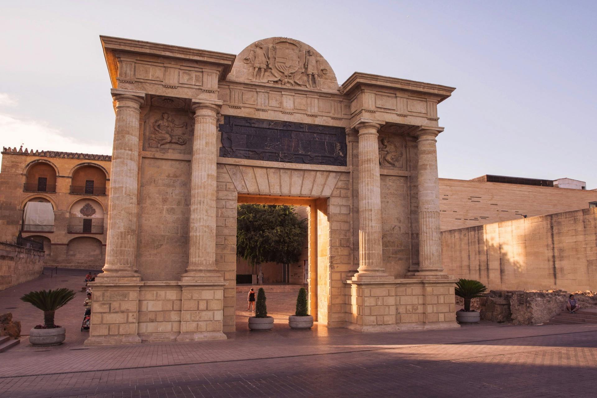 Stone archway with ornate carvings and columns, leading to a courtyard with trees. Two people walking through the arch.