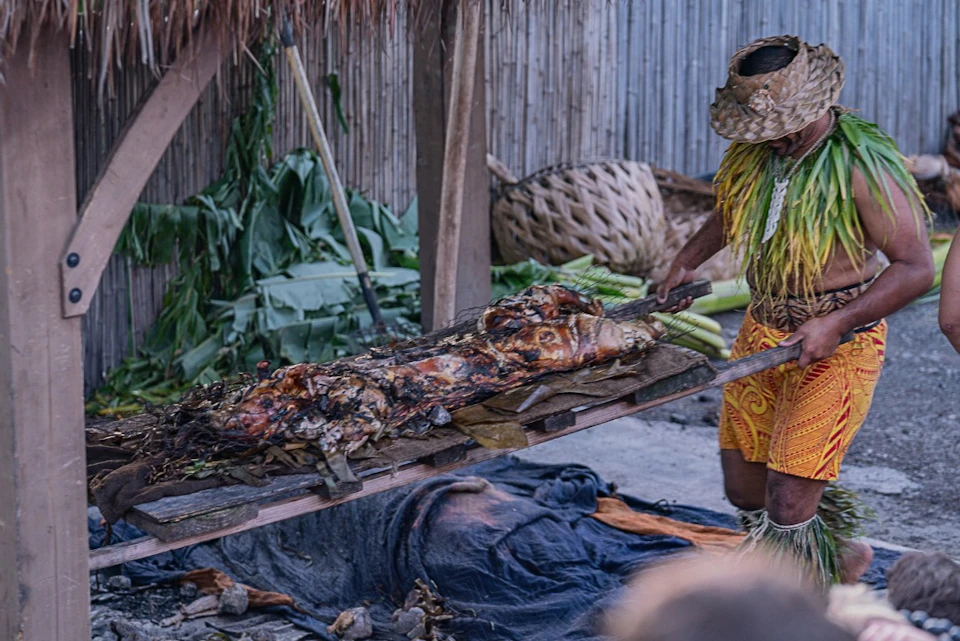 A person wearing traditional attire holds a tray with a roasted animal, surrounded by leaves and a bamboo fence in the background.
