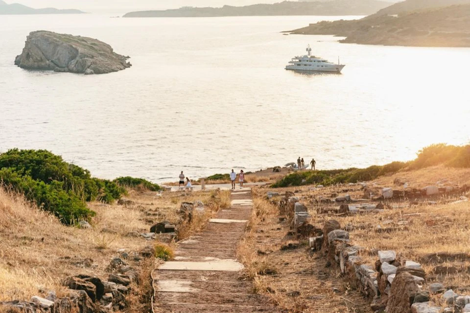 A coastal scene with a yacht on the water, a rocky path leading down through dry vegetation, and people walking towards the shore.
