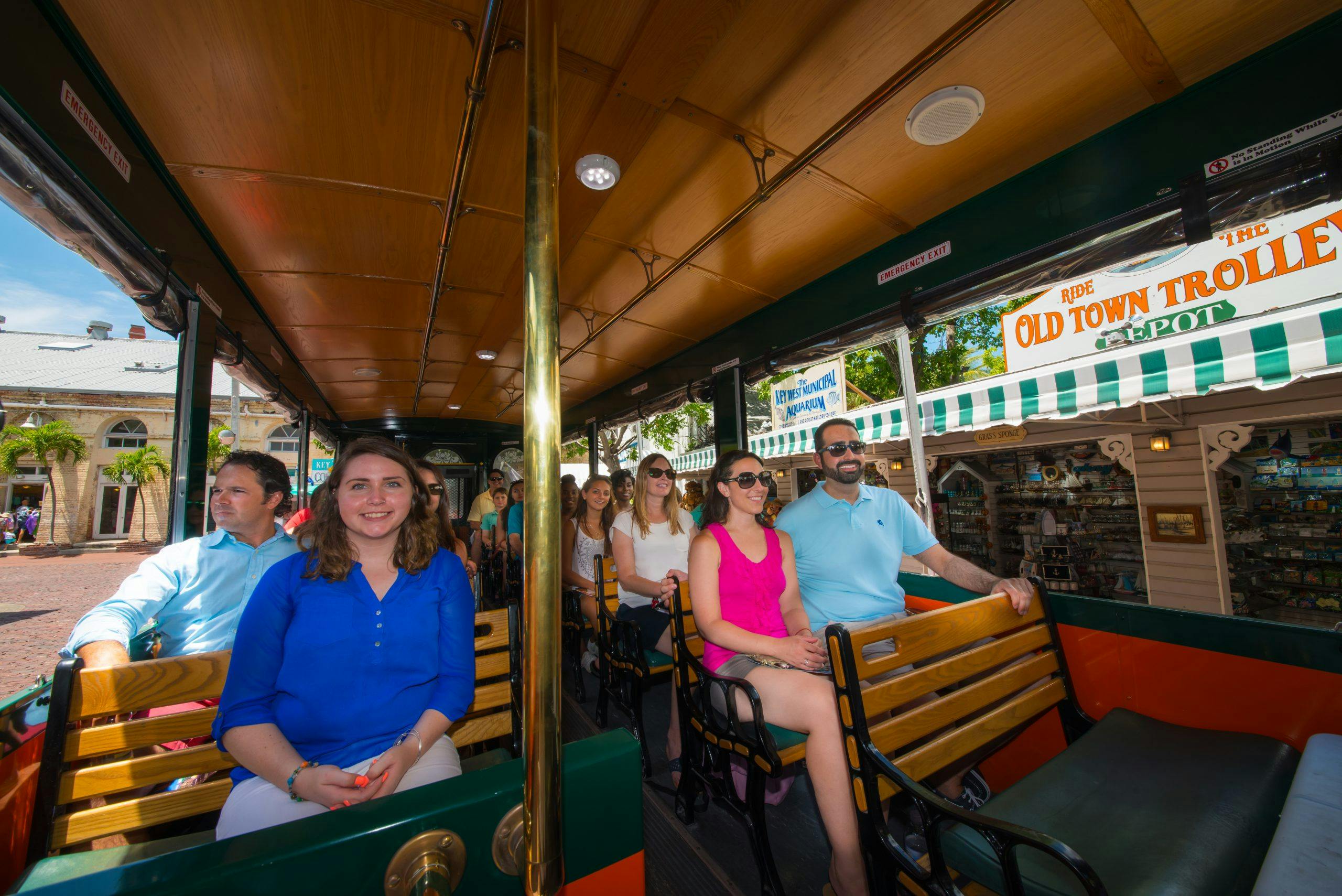 Skupina lidí sedí uvnitř venkovního vozíku. Žena v modré košili se usmívá do kamery, ostatní se dívají před sebe. Slunečný den venku.