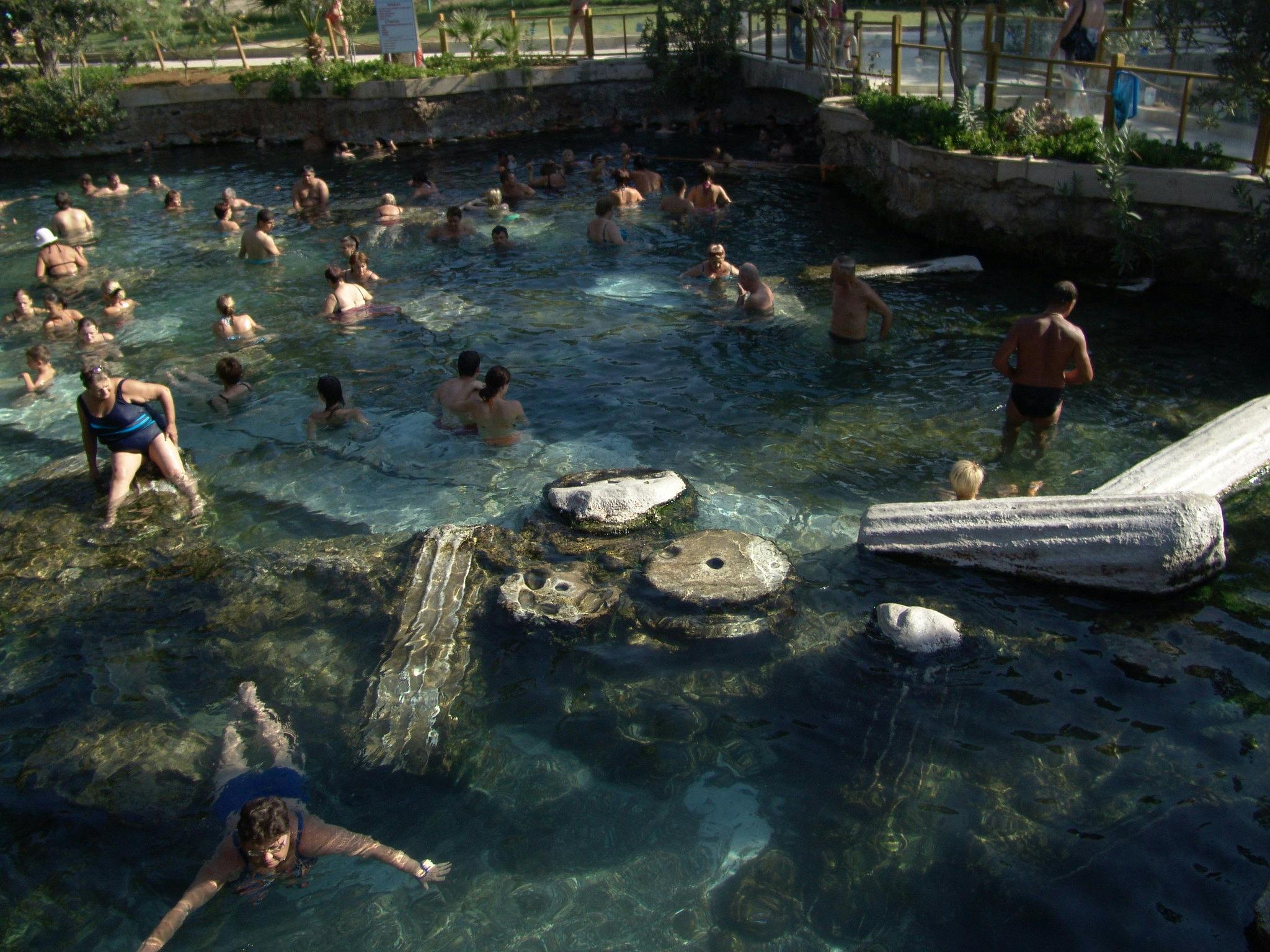 古代の柱や石が部分的に水没した天然プールで泳ぐ人々。 このエリアは緑に囲まれています。