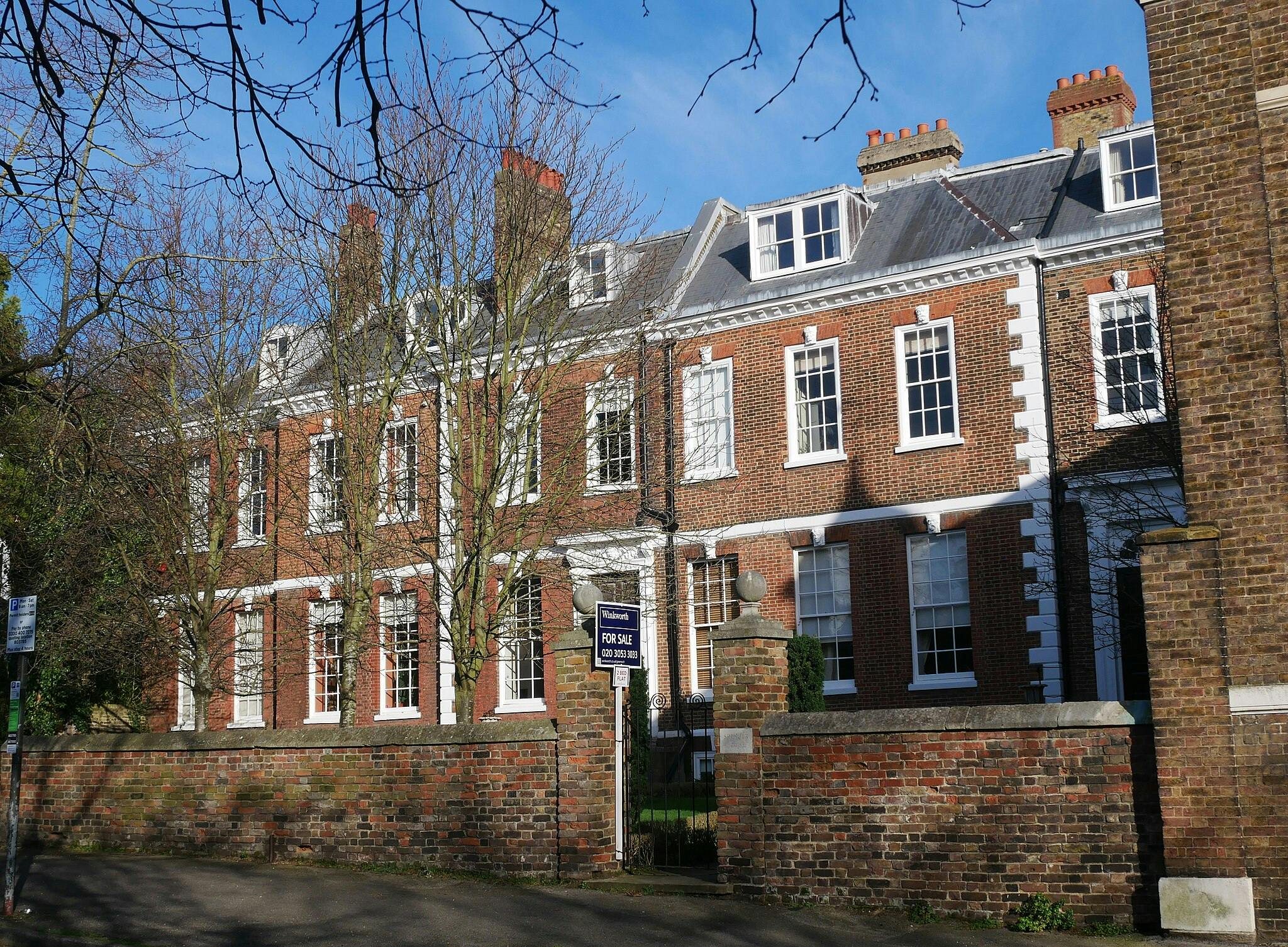 도머 창문과 굴뚝이 있는 붉은 벽돌 주택이 줄지어 있고, 앞에 '매매' 표지판이 있으며, 벽돌 벽을 따라 잎이 없는 나무가 있습니다.