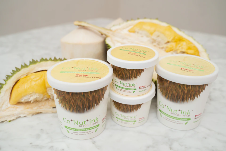 Four containers of Co+Nut+ink durian-flavored coconut ice cream on a marble surface next to fresh durian and a coconut.