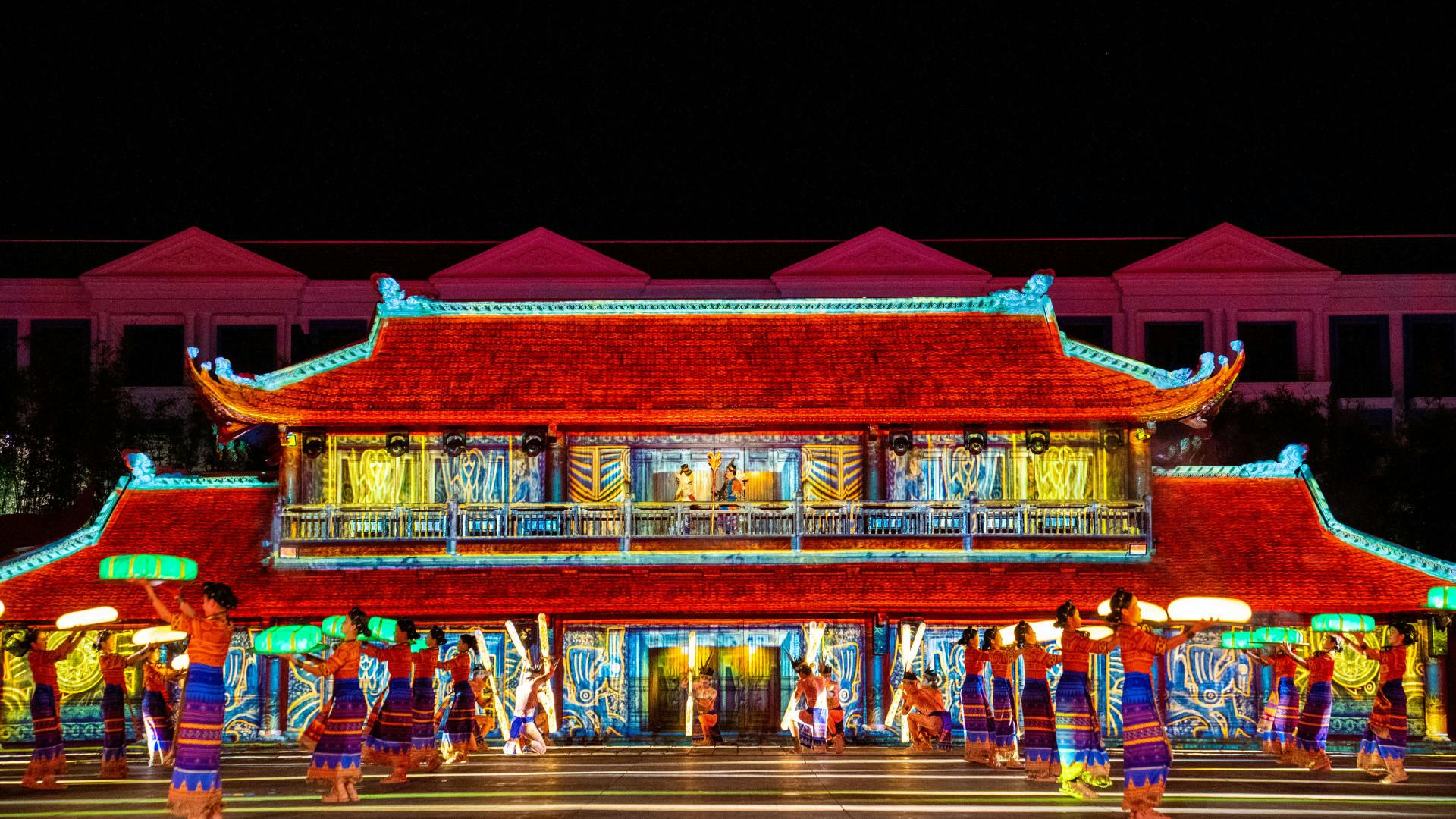 A brightly illuminated traditional-style building with colorful projections and performers in vibrant costumes in front.