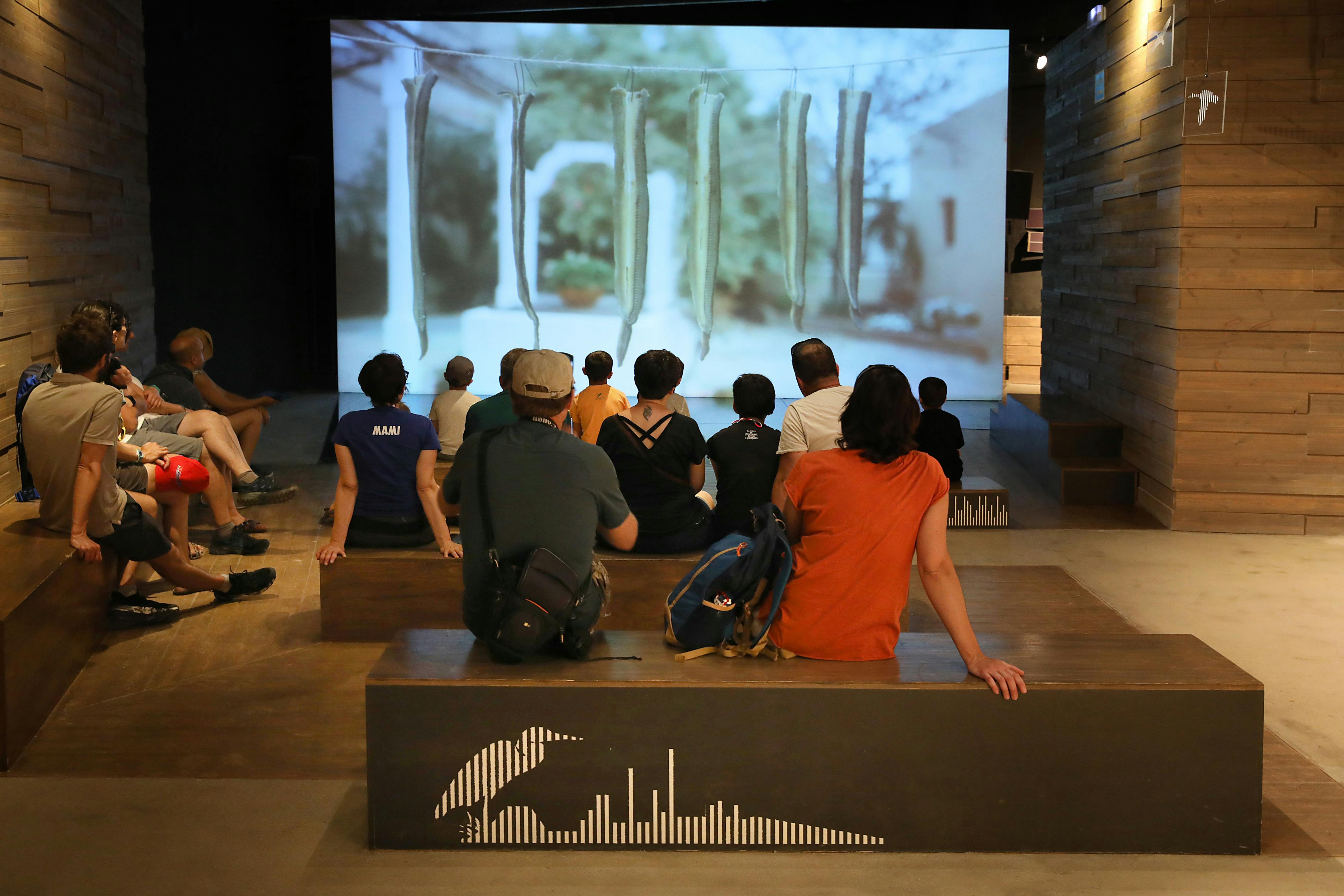 Άνθρωποι κάθονται σε ξύλινα παγκάκια μέσα σε μια γκαλερί, παρακολουθώντας μια μεγάλη οθόνη που εμφανίζει κρεμαστά αντικείμενα έξω από ένα κτίριο.