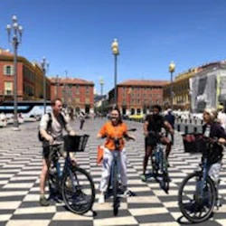 Fyra personer med cyklar som står på ett svartvitt rutigt torg, omgivet av färgglada byggnader och en klarblå himmel.