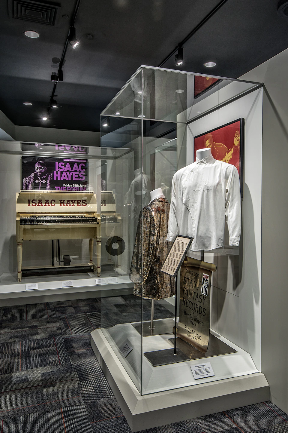 Museum display of Isaac Hayes memorabilia, including clothing, a piano with his name, and framed art.