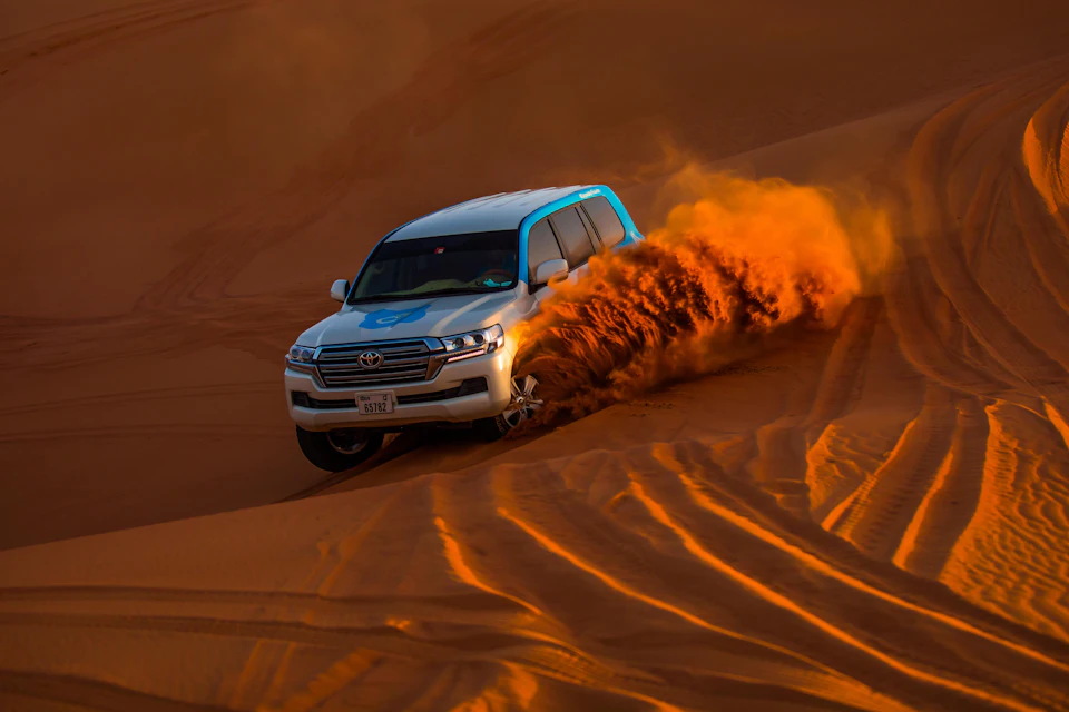 A white SUV with blue accents drives through sand dunes, kicking up a cloud of orange sand in the desert.