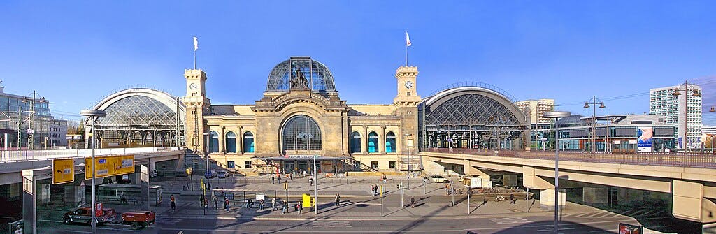 大型火车站，拱形玻璃屋顶，中央钟楼，人们步行，多栋建筑，背景是晴朗的蓝天。