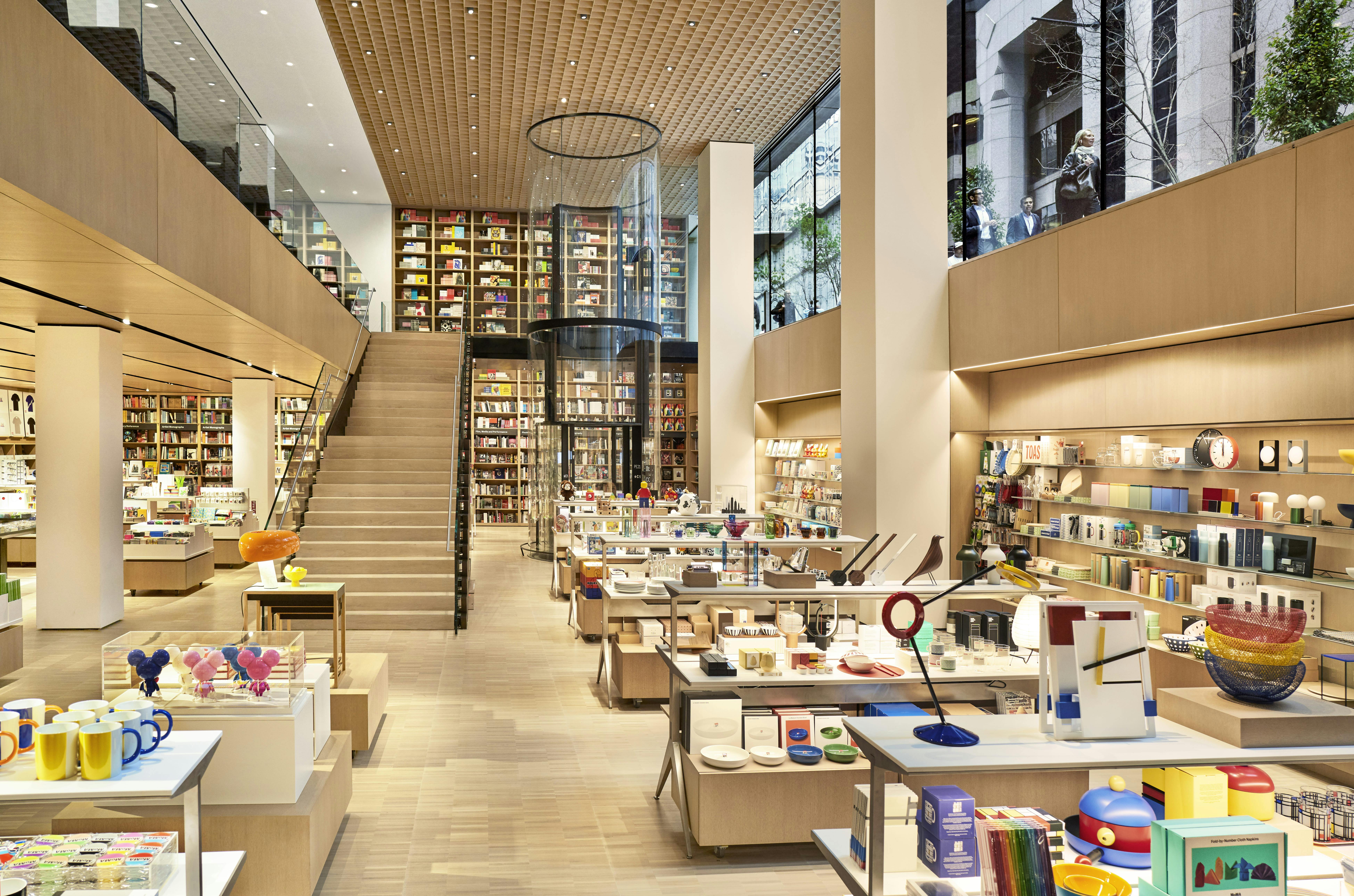 Dvoupatrové moderní knihkupectví s dřevěnými policemi, různorodým zbožím, centrálním schodištěm a velkými skleněnými okny.