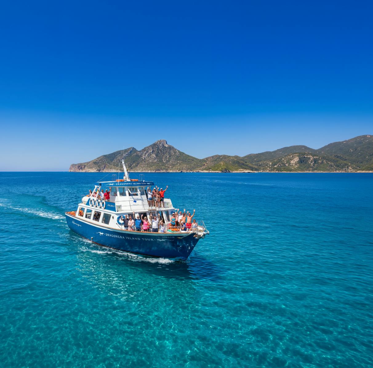 Un tour in barca blu con passeggeri che navigano su acque limpide e turchesi, con una costa montuosa sullo sfondo sotto un cielo blu.