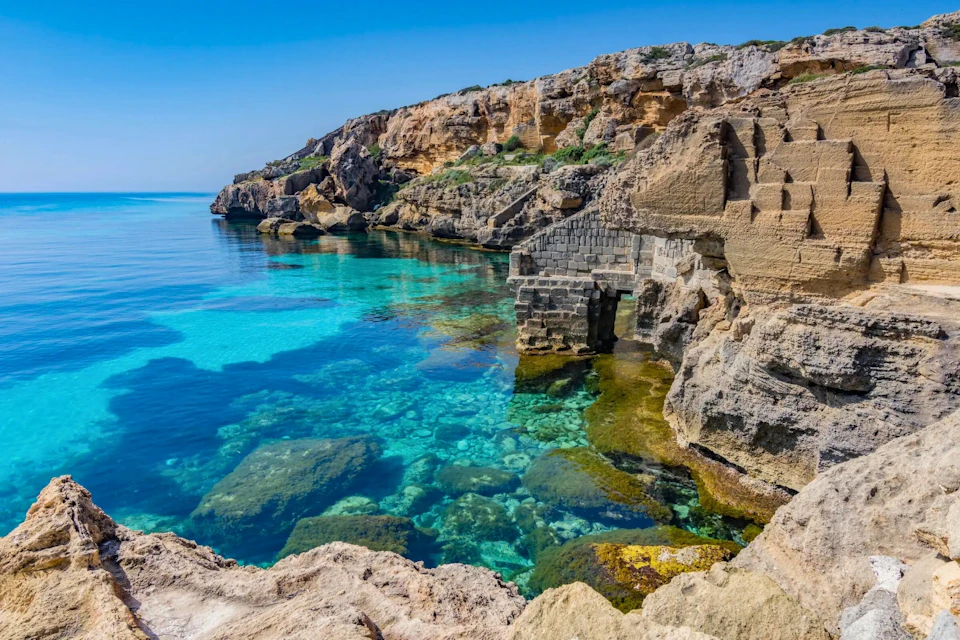 A rocky coastline with clear, turquoise water and a stone structure near the shore under a bright blue sky.