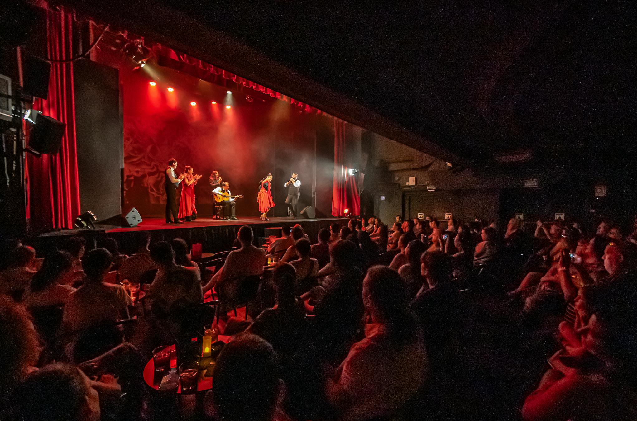 Živé vystoupení na jevišti s tanečníky v červených šatech a hudebníky, při pohledu publika v tlumeně osvětleném divadle.