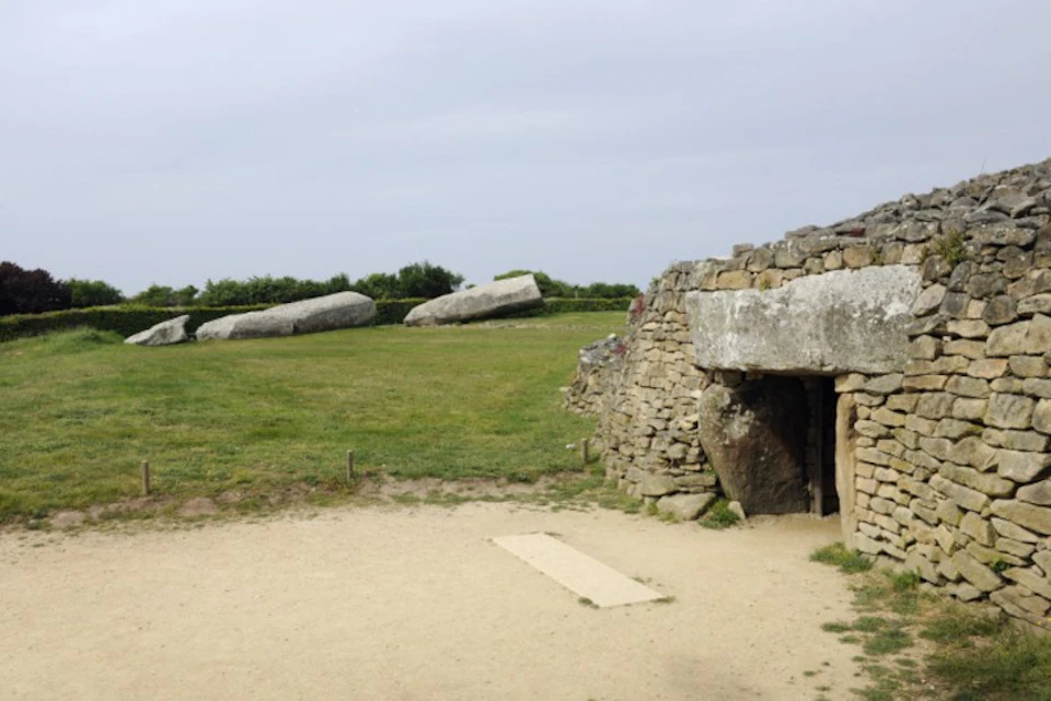 Stone structure with a grassy field containing large rock formations in the background, under a cloudy sky.