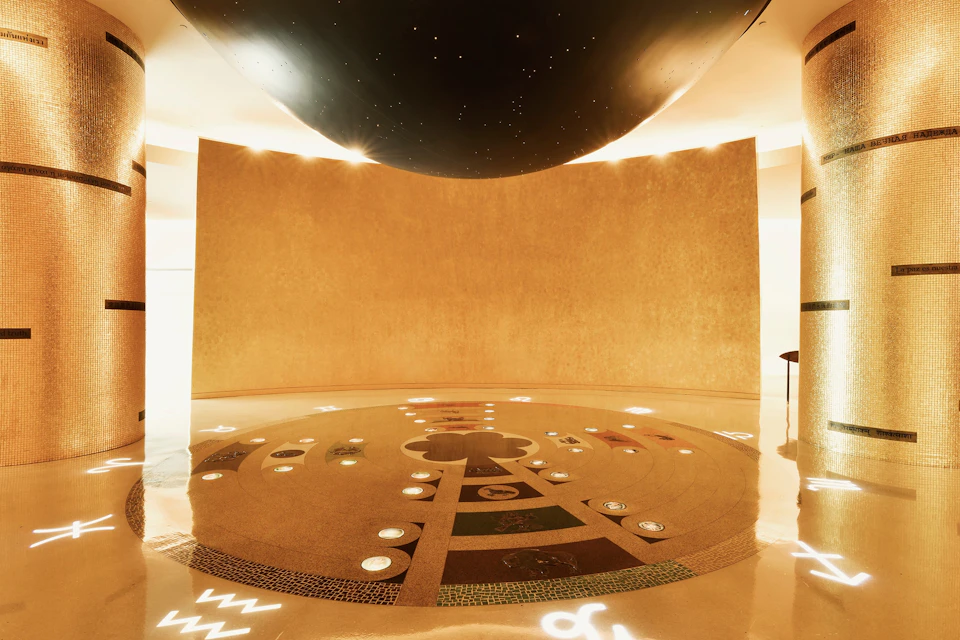A circular, illuminated design on the floor with symbols, under a black dome with star-like lights and beige walls.