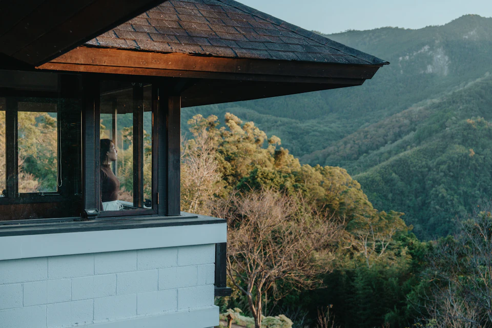 A person sits inside a building with large windows, overlooking a scenic view of trees and forested hills.