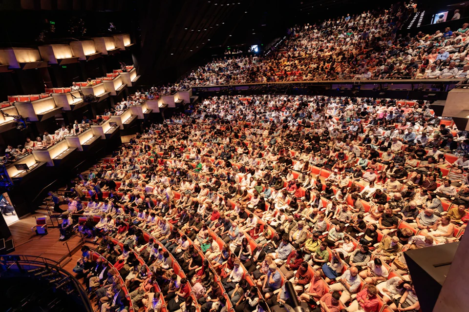 A large audience seated in a multi-tiered theater with rows of red seats and balcony sections under dim lighting.