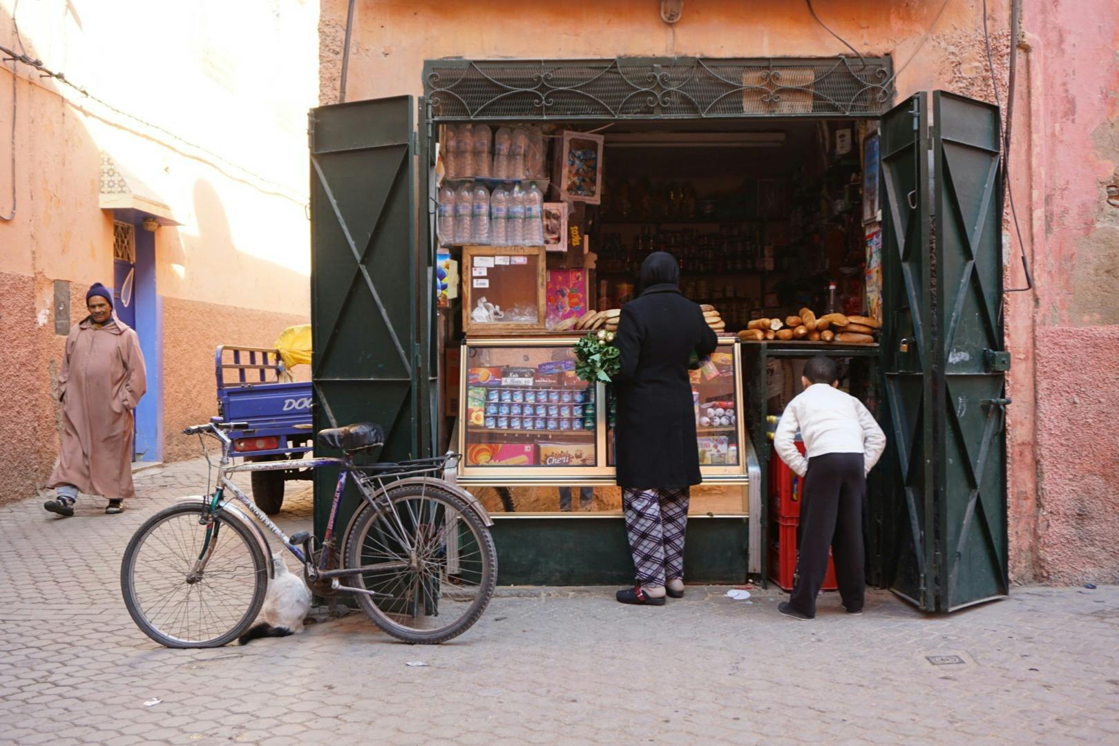 A woman in a black coat stands at an outdoor market stall with a bicycle and a blue cart nearby. Another person is seen walking.
