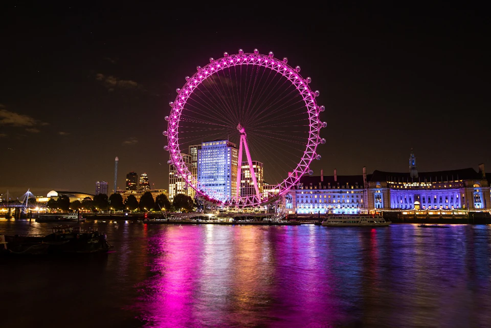 A brightly illuminated Ferris wheel with pink lights, set against a city skyline at night, reflecting on the water below.