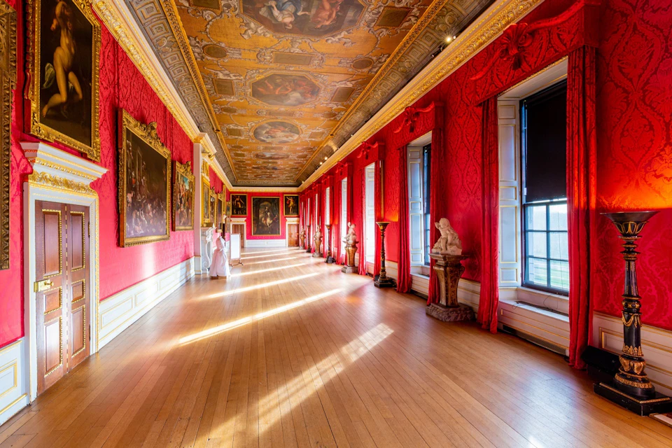 A richly decorated gallery with red walls, a painted ceiling, large windows, statues, and framed artworks along the walls.