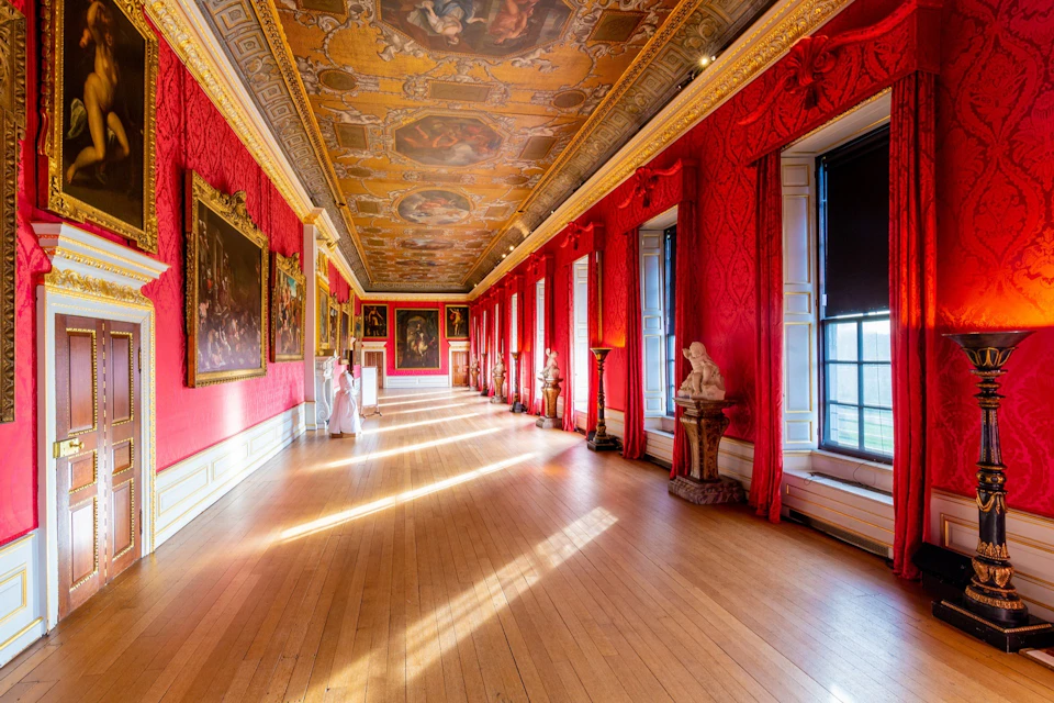 A richly decorated gallery with red walls, a painted ceiling, large windows, statues, and framed artworks along the walls.