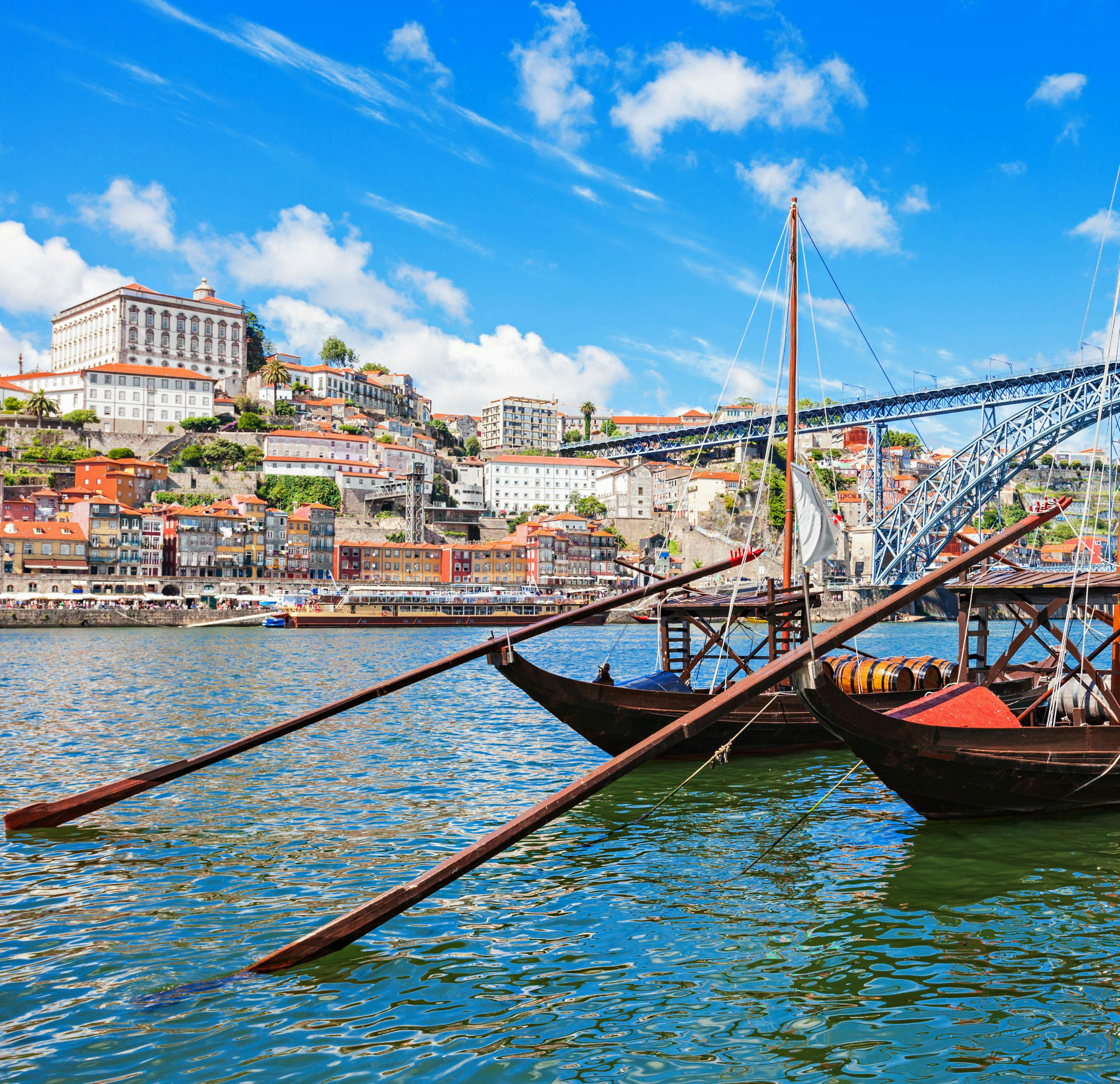 Lodě s barely kotvící na řece s výhledem na panorama města s barevnými budovami a velkým mostem pod modrou oblohou.