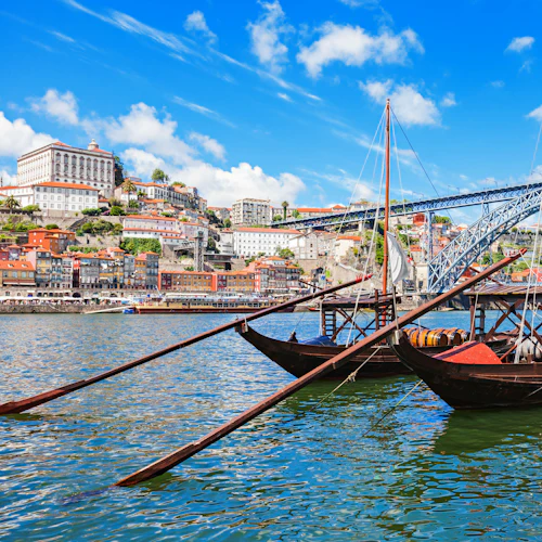 Båtar med tunnor dockade på en flod, med utsikt över en stadsbild med färgglada byggnader och en stor bro under en blå himmel.