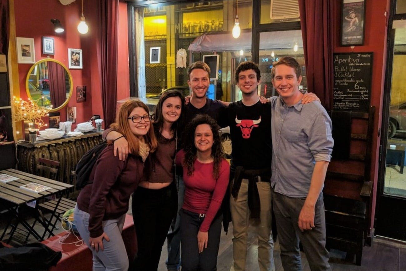 Five people are smiling and standing together in a bar or café, with red curtains and a window in the background.