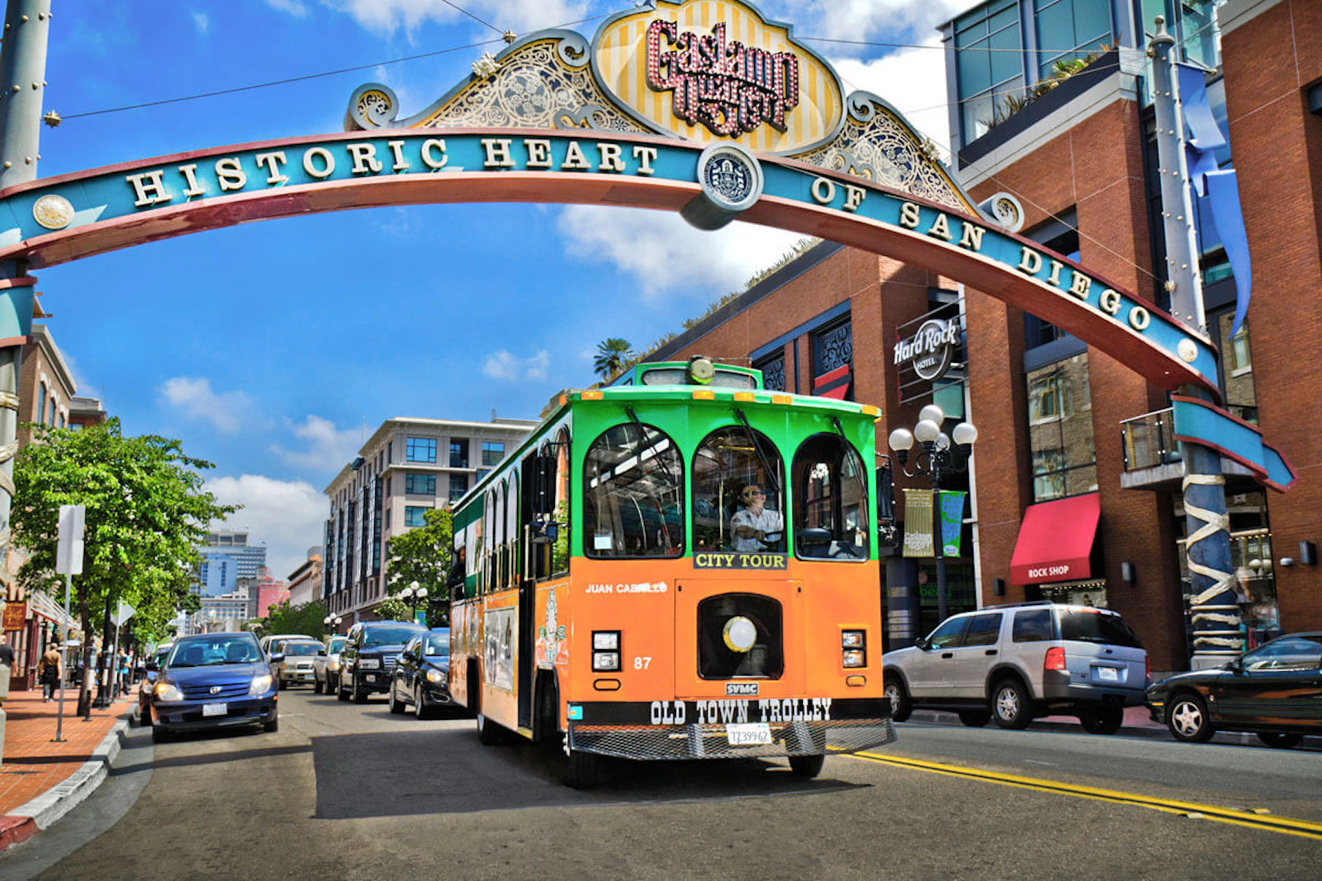 An orange and green trolley bus on a street under the "Historic Heart of San Diego" arch with buildings and cars in the background.