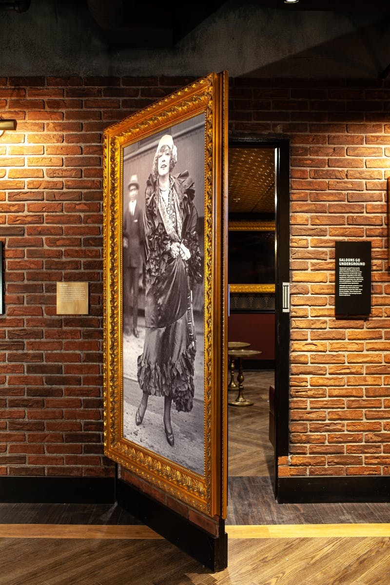 A hallway with brick walls features a large vintage photo of a woman in an elegant dress on a door, leading to an interior room.