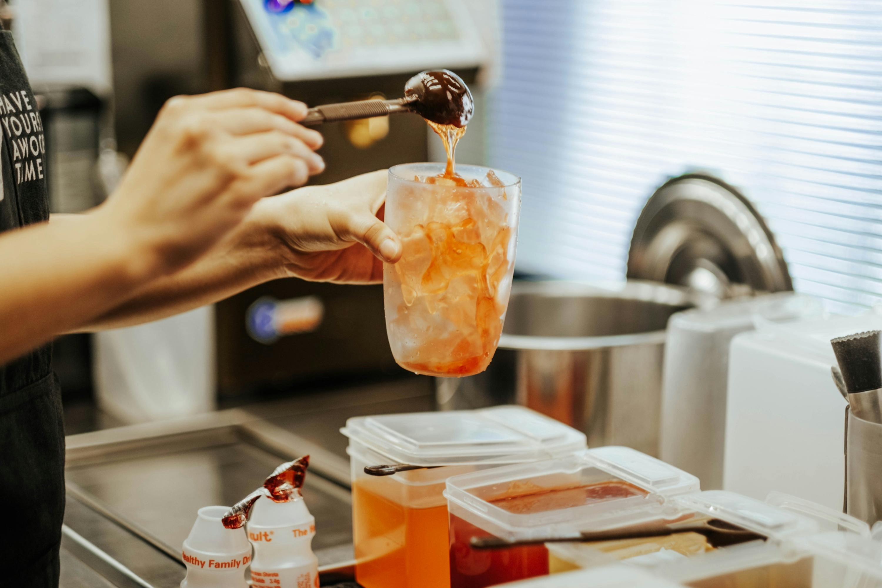 A person preparing a drink by adding syrup from a spoon into a plastic cup filled with ice, in a kitchen setting.