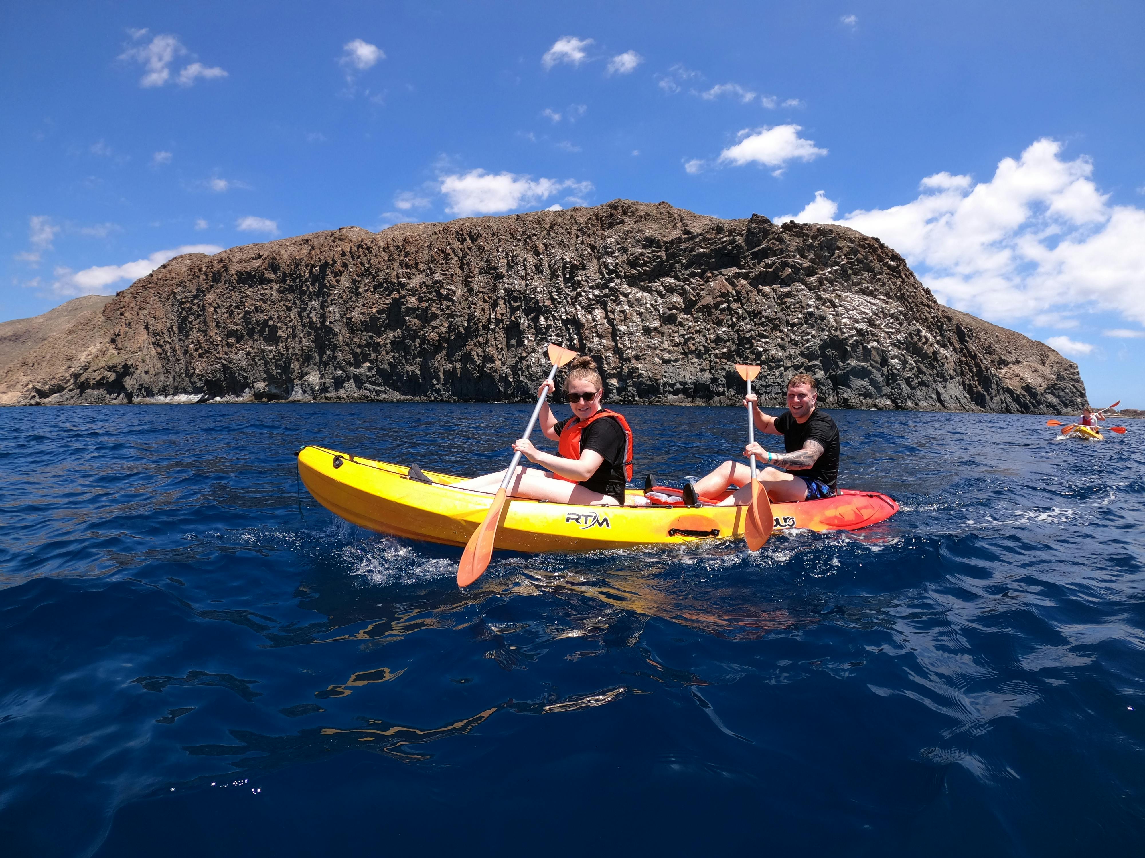 两个人在晴朗的蓝天下划着一艘黄色皮划艇，后面是一个岩石悬崖。