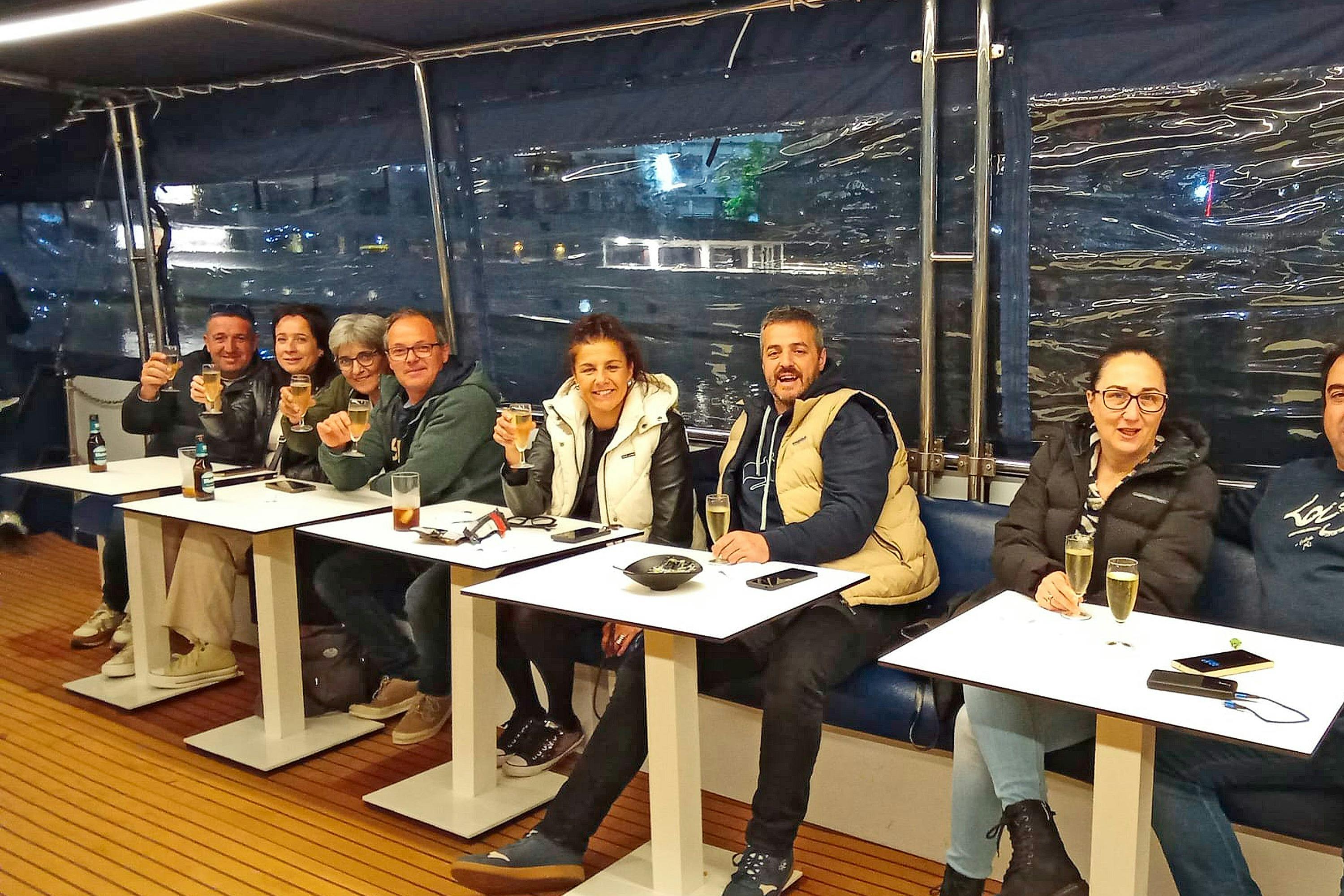 A group of people sitting at tables indoors, smiling and holding glasses, with a nighttime setting visible outside.