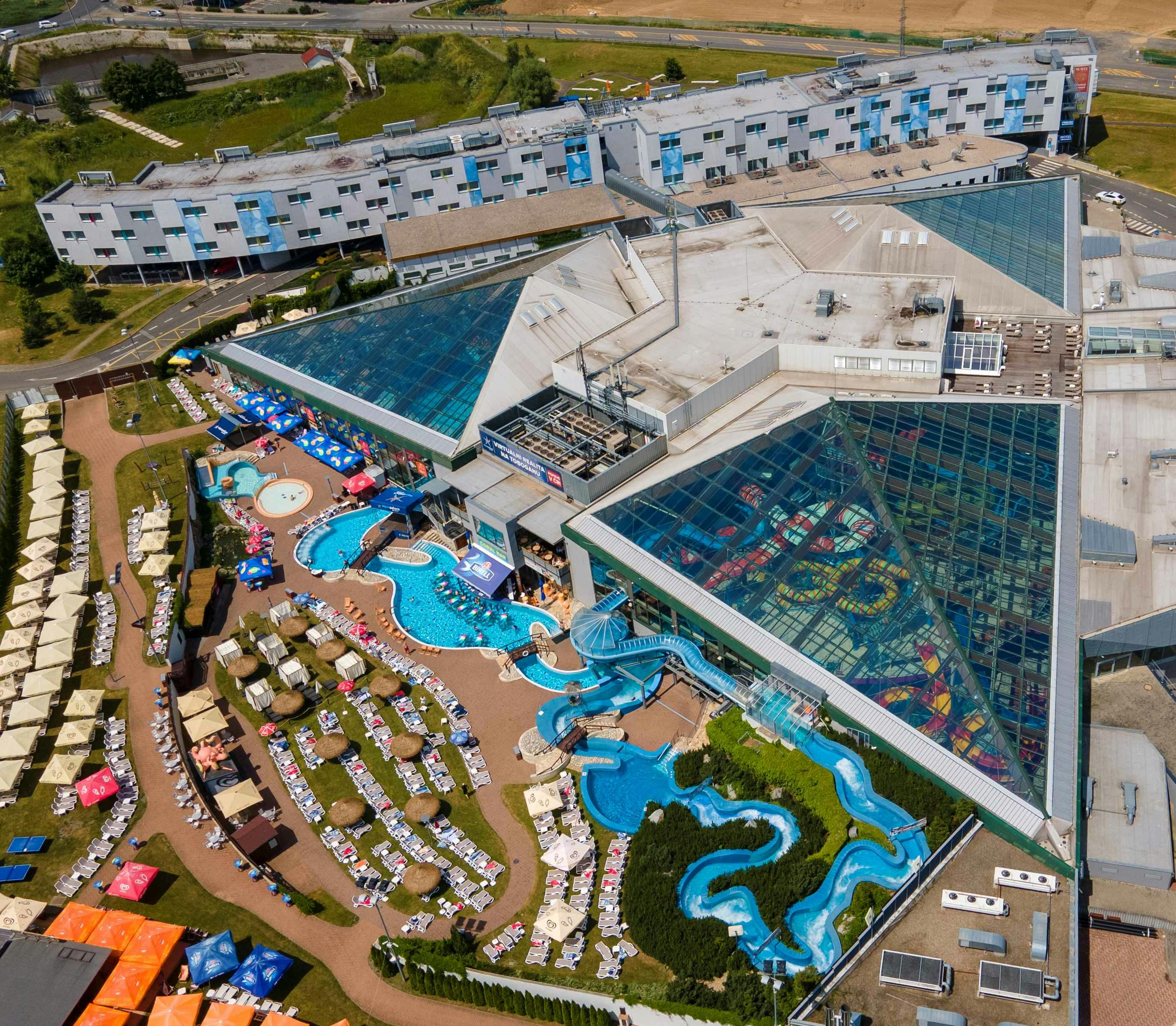 Aerial view of a large aquatic complex with outdoor pools, water slides, sunbathing areas, and a glass-roofed building.
